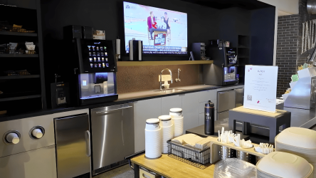 Self-serve drink and condiment station with coffee machines, cups, straws, lids, and a large TV mounted above a counter in a cafeteria or lounge area.