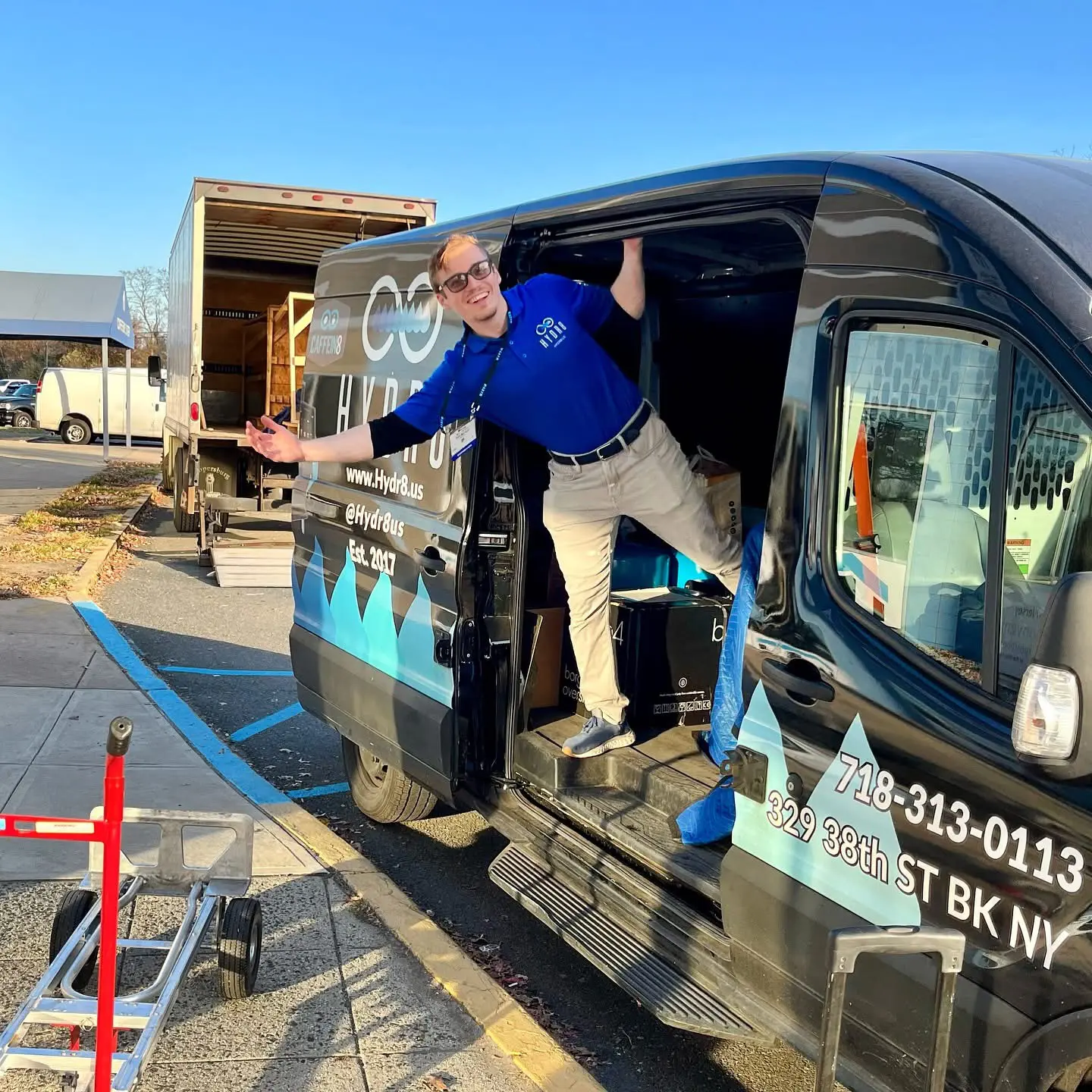 A person wearing a blue shirt and khaki pants poses cheerfully in the doorway of a black van parked in a lot, with equipment and a hand truck nearby.