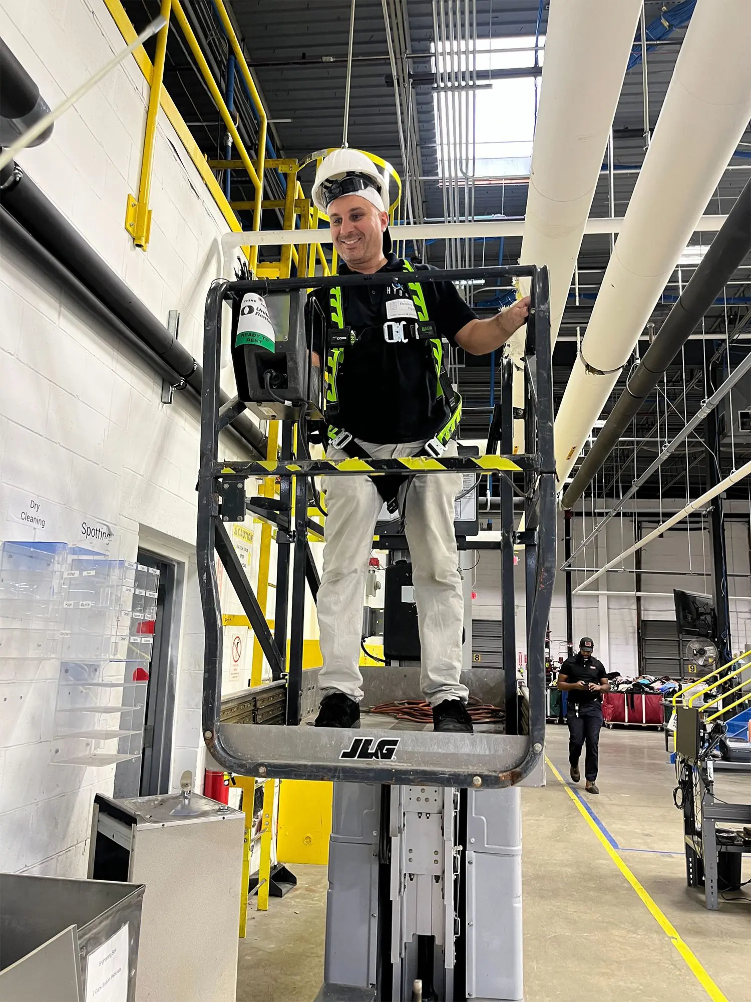 A worker wearing safety gear stands on a raised JLG lift inside an industrial facility with pipes and equipment visible in the background.