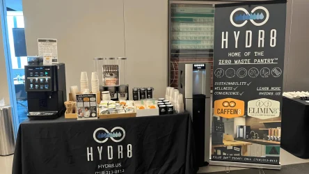 A self-serve drink station with a coffee machine, cups, condiments, and a HYDR8 banner promoting zero waste and sustainable beverage options.